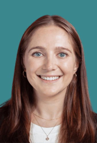 A woman with long brown hair and blue eyes smiles at the camera, wearing gold hoop earrings and layered necklaces, against a teal background.